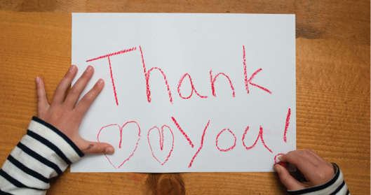 两个孩子的手用红色蜡笔在一张白纸上画着“谢谢你”和两颗心