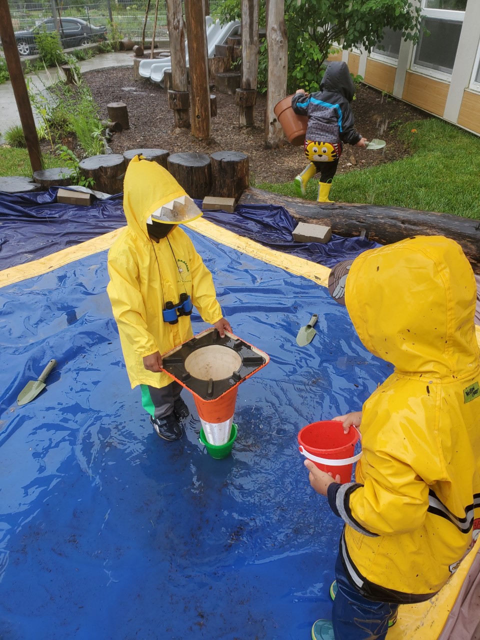 两个穿着雨衣和靴子的孩子在外面玩一个倒置的交通锥和水桶