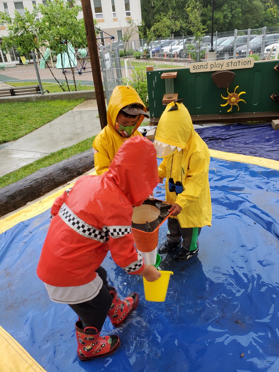 三个穿着雨衣和靴子的孩子玩着水桶、水和一个倒置的交通锥