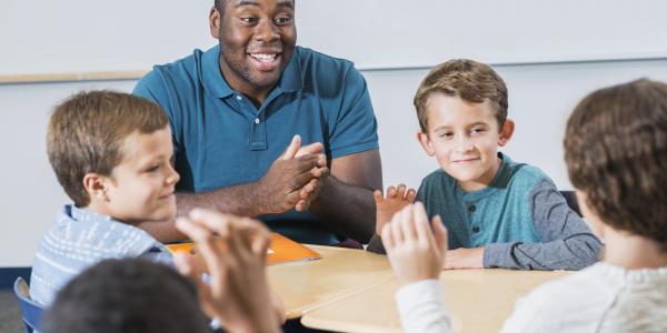 年轻的有色人种黑人男性教师与一群白人男孩坐在一张桌子旁。