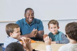 年轻的有色人种黑人男性教师与一群白人男孩坐在一张桌子旁。
